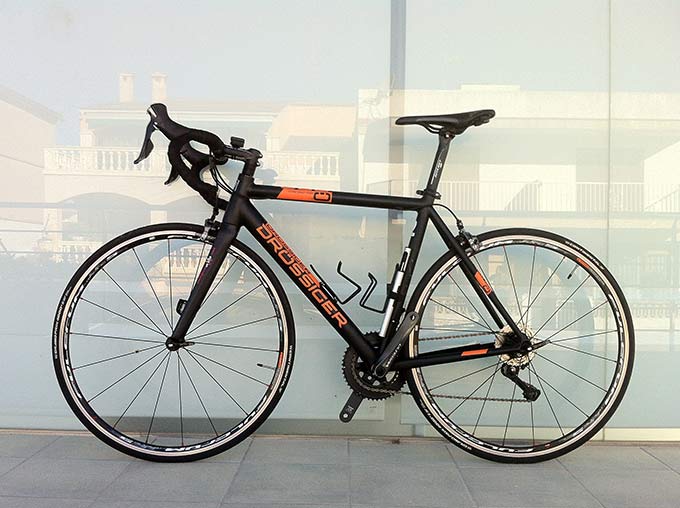 Black and orange Drossiger road bike leaning against a glass panel, drop handlebars and drivetrain visible
